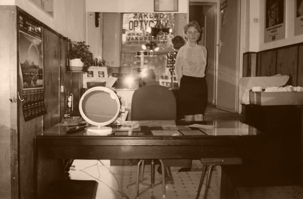 Historyczne zdjęcie z salonu optycznego Jakubowicz przy ul. Marymonckiej 63 w Warszawie – trzy osoby we wnętrzu pracowni: młoda kobieta, starsza kobieta i mężczyzna w białym fartuchu. W tle witryna z oprawkami i klasyczny wystrój. Od lat specjalizujemy się tu w doborze okularów progresywnych.