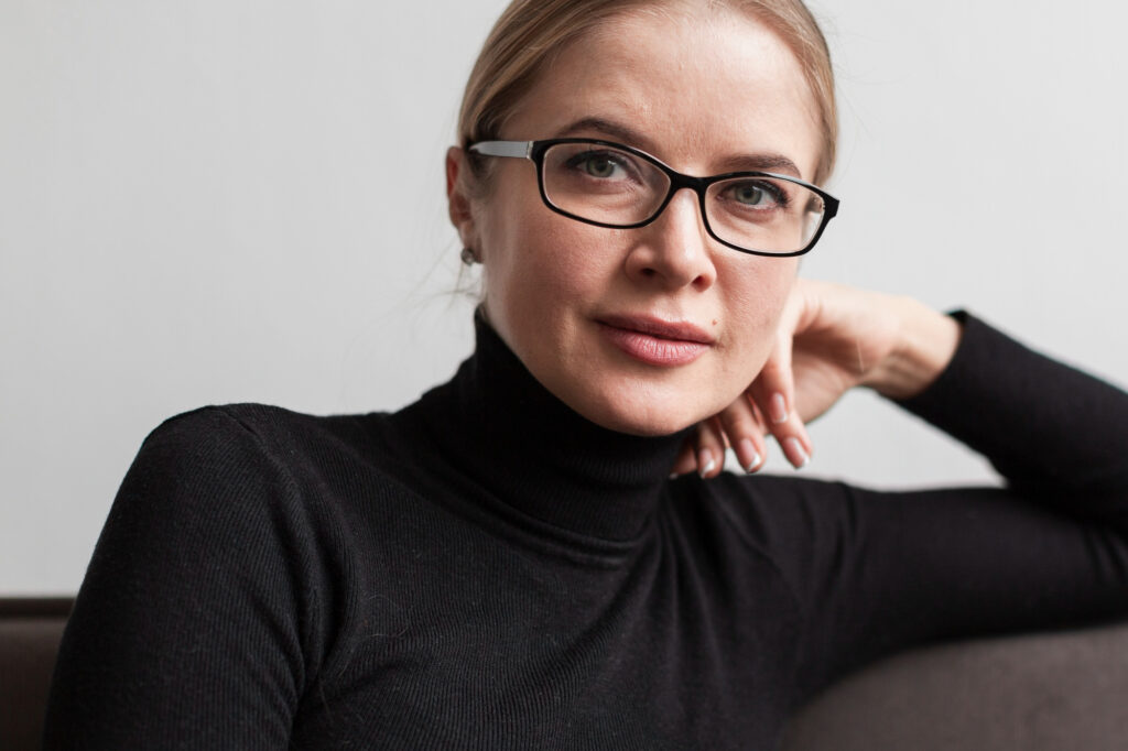 Kobieta w okularach progresywnych Varilux, siedząca w czarnym golfie, z lekkim uśmiechem, wsparta na dłoni – naturalny portret w spokojnym, minimalistycznym stylu.
