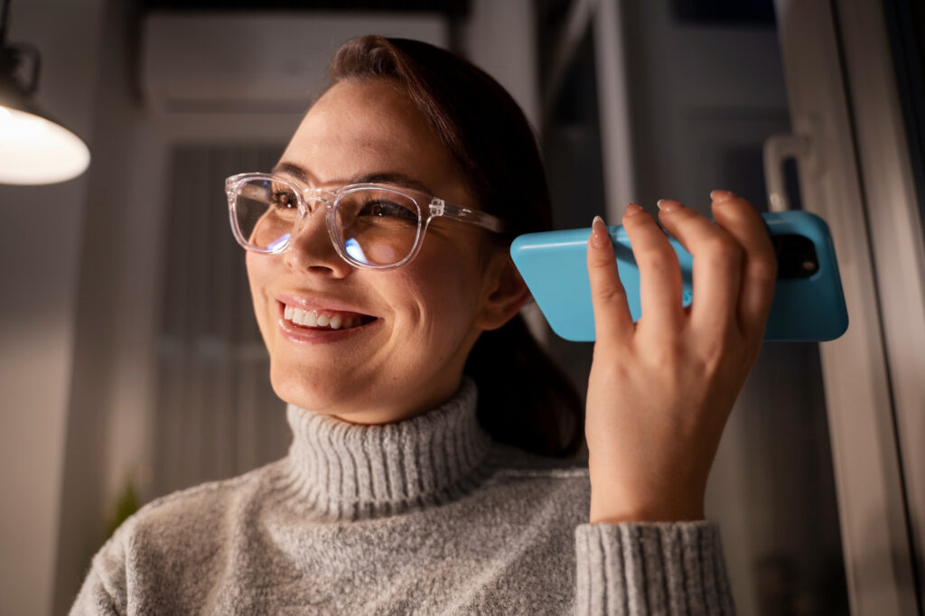 Uśmiechnięta kobieta rozmawiająca przez telefon, w okularach ze szkłami z antyrefleksem – wieczorne światło odbija się subtelnie od szkieł.