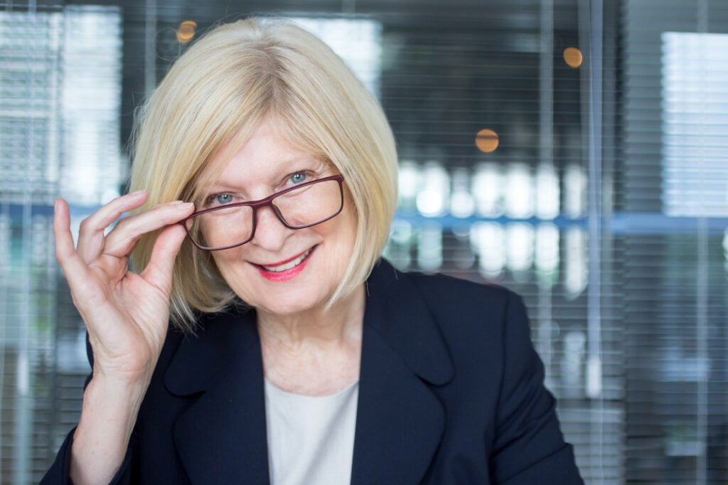 Kobieta w średnim wieku w okularach do pracy biurowej od Salonu Optycznego Jakubowicz, uśmiecha się i spogląda znad oprawek w nowoczesnym biurze – symbol komfortu widzenia i profesjonalizmu.