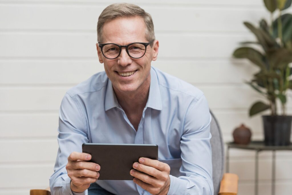 Uśmiechnięty mężczyzna w okularach progresywnych Hoyalux siedzi wygodnie w fotelu, trzymając tablet – cieszy się wyraźnym widzeniem na każdą odległość i nowoczesnym designem oprawek.